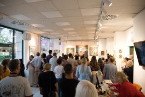 A large crowd of attendees gathered inside the well-lit Golden Duck Gallery for the opening reception of an international art exhibition.